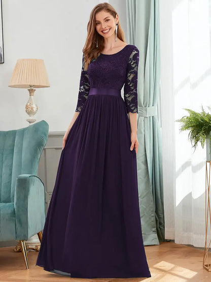 Woman wearing a long purple dress with lace sleeves in a room with a lamp and plant.