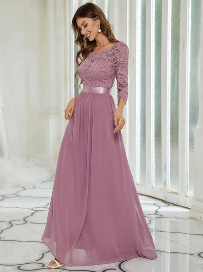 Woman wearing a long, elegant purple dress with lace top and flowing skirt in a bright room.