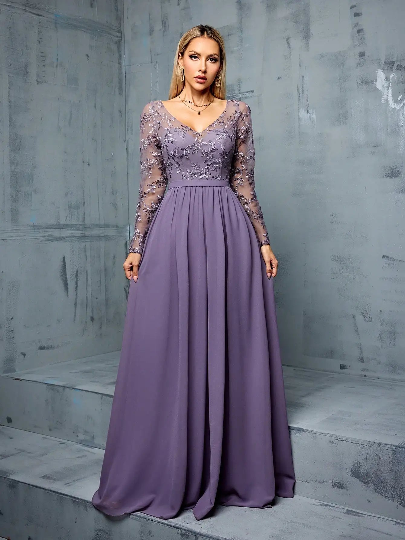 Woman wearing a long purple dress with lace sleeves against a gray concrete wall.