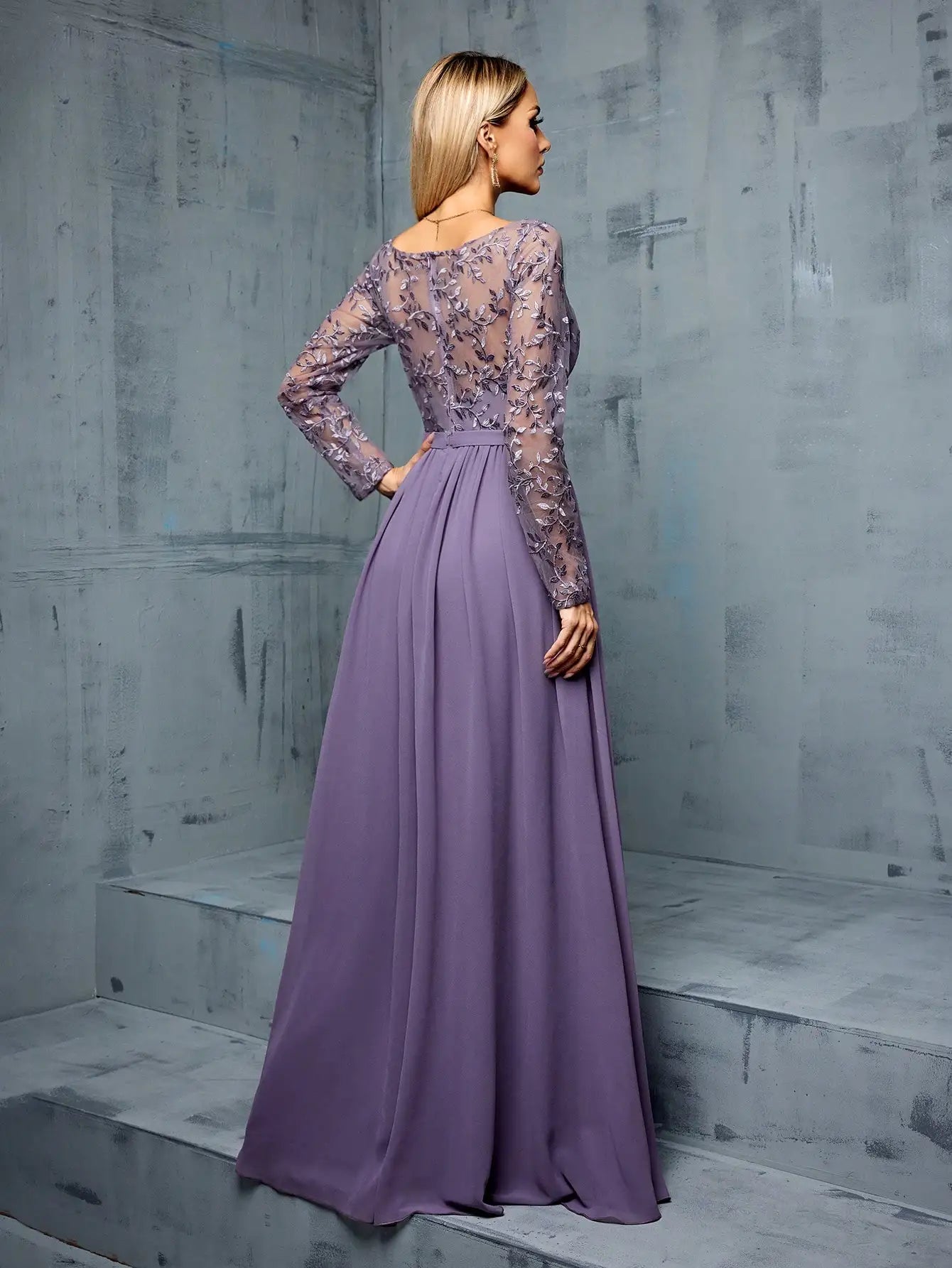 Woman wearing a long purple dress with lace detailing against a gray concrete wall.